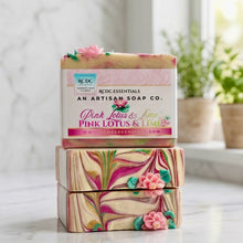 Three stacked bars of handmade soap from Redc Essentials, featuring a pink and lime scented bar on top with a paper label, and two marbled bars below with pink lotus flowers, set on a marble countertop with a blurred kitchen background.