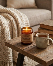 A lit amber glass jar candle labeled 'Fall Into Cory' sits on a rustic wooden table beside a ceramic mug and stacked books, with a cozy knitted throw and sofa in the background.
