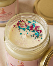 An open glass jar candle filled with creamy off-white wax and topped with colorful glitter, shown with its gold lid beside it on a light neutral surface.