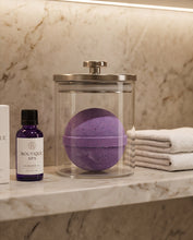 A purple bath bomb sits inside a clear glass jar with a silver lid on a marble countertop, with a Boutique Spa lavender oil bottle and folded white towels nearby.