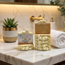 Two decorative bars of soap displayed on a marble bathroom counter, with a rolled towel, small potted succulent, and warm vanity lighting in the background.