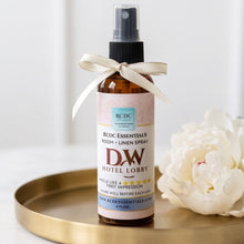 A bottle of WD Hotel Lobby Room + Linen Spray by RCDC Essentials sits on a round gold tray next to white flowers. The amber glass bottle has a black spray top and is tied with a cream ribbon.