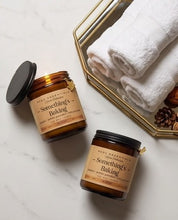 Two amber glass jars of ACDF Essentials 'Something's Baking' candles are arranged on a white marble surface next to a gold-framed tray holding white towels.