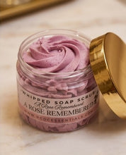 A clear jar of whipped soap scrub with a gold lid leaning against it on a light marble surface. The scrub is a dusty mauve-purple color, and the label reads 'Whipped Soap Scrub A Rose Remembered' with the brand 'BCDCEssentials'.