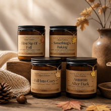Four amber glass soy wax candles in black metal jars with kraft paper labels from HCCD Essentials Home Collection, styled on a rustic wooden surface with autumn leaves, a knitted blanket, and a ceramic vase in the background.