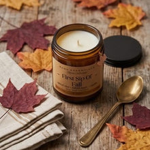 An amber glass jar candle labeled 'First Sip Of Fall' by Wood Essentials, styled on a rustic wooden surface with scattered autumn leaves, a folded cream napkin, and a gold spoon.