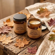 Two amber glass candles labeled 'Autumn After Dark' by The Candle Company, styled on a rustic wooden table with scattered autumn leaves and a cream knit blanket.