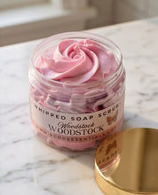 A clear jar of pink whipped soap scrub with a white label reading 'Woodstock Woodstock' and a gold lid resting beside it on a light marble surface.
