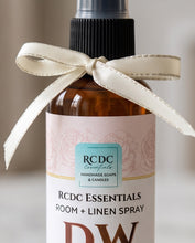 An amber glass spray bottle of RCDC Essentials Room + Linen Spray, decorated with a cream satin ribbon tied in a bow around the neck, set against a soft, blurred neutral background.