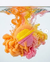 A colorful bath bomb or sponge product dissolving in water, creating vibrant orange, yellow, and pink swirls against a clean white background.