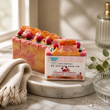 A photo of bars of handmade soap decorated with raspberry and tangerine designs, with a slice of the bar showing the swirled pink and white colors. The soap is accompanied by decorative flowers and the packaging is visible.