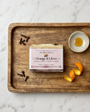 A packaged bar of soap labeled 'Orange & Clove' by ACDC ESSENTIALS, placed on a wooden tray with orange peel, cloves, and a small dish of honey on a white marble surface.