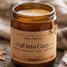 An amber glass jar candle with a kraft paper label reading 'RCDC ESSENTIALS Home Collection - Fall Into Cozy -' and a small gold leaf charm, set on a rustic wooden surface with soft neutral fabric in the background.