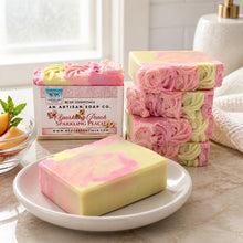 A bar of handmade cold process goat's milk soap with pink and yellow layers, decorated with a floral design, next to a vase with peach flowers.