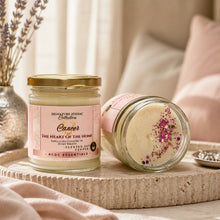 Two scented candles from the Signature Zodiac Collection 'Cancer' blend are displayed on a stone tray. One candle is upright in a clear glass jar with a gold lid and a pink label, while the other lies on its side showing a creamy wax surface decorated with pink and purple dried flower petals.