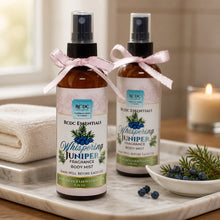 Two bottles of RCDC Essentials Whispering Juniper Fragrance Body Mist on a marble surface with a candle and dish of juniper berries in the background.