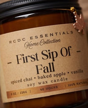 An amber glass soy wax candle jar with a black lid and a kraft paper label reading 'RCDC ESSENTIALS Home Collection - First Sip Of Fall' with scent notes of spiced chai, baked apple, and vanilla.