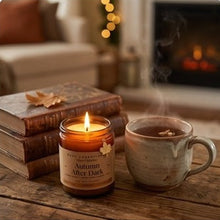 A lit candle in a glass jar labeled 'Autumn After Dark' sits on a wooden table beside a steaming ceramic mug, with stacked books and a cozy fireplace in the background.