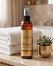 An amber glass room and linen spray bottle with a gold label from RCDC Essentials Home Collection, styled on a marble bathroom counter next to folded white towels and a small succulent in a gold pot.