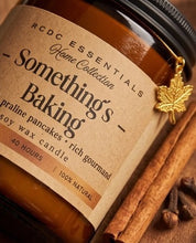 A close-up of an RCDC Essentials Home Collection soy wax candle in a brown glass jar, labeled 'Something's Baking' with a kraft paper label and a small gold maple leaf charm, styled with cinnamon sticks and cloves on a warm brown background.