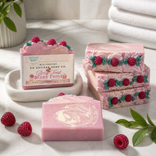 Pink raspberry twist artisan soap bars arranged on a white marble surface with fresh raspberries and green leaves. One soap bar is shown in its branded retail box, with additional stacked soap bars beside it.