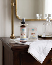 A bottle of DW Hotel Lobby room freshener and a boxed matching room freshener sit on a white cloth draped over a dark wood dresser, with a brass lamp and glass decanter in the background.