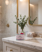 A boxed tea product labeled 'English Tea Garden' sits on a white marble bathroom counter beside a glass vase with eucalyptus stems, folded towels, and a gold-framed mirror in a bright bathroom setting.