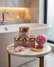 Three pink and gold jars of whipped soap are arranged on a round marble side table in a bright bathroom setting, with a matching pink soap bar and flower petals nearby.