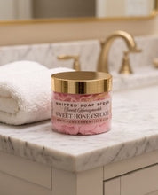 A jar of KCDG Essentials Sweet Honeysuckle Whipped Soap Scrub sits on a white marble bathroom counter next to a folded white towel, with a gold faucet and mirror in the background.