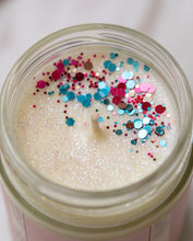 A close-up top-down view of a clear glass jar candle filled with creamy off-white wax and topped with colorful glitter confetti in pink, blue, red, and silver.
