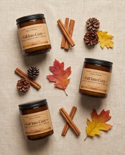 Three amber glass candles with black lids and tan labels reading 'Fall Into Cozy' are arranged on a light beige fabric background with cinnamon sticks, pinecones, and autumn leaves.