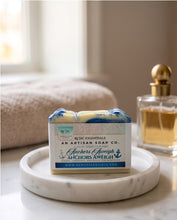 A bar of handmade soap in a white paper box with blue and green accents, placed on a round white marble tray. A perfume bottle is visible in the background on a bathroom counter.