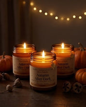 Three lit amber glass candles labeled 'Autumn After Dark' by SCDC Essentials, arranged on a dark wooden surface with small pumpkins and dried botanicals, under warm string lights.