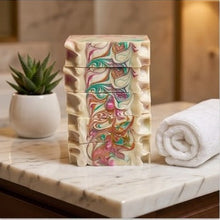 A stack of handmade rectangular bars of soap with a cream base and colorful marbled swirls of pink, green, orange, and blue, displayed on a marble countertop next to a rolled white towel and a small potted succulent.