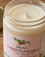 A close-up of a Poma's 'Garden Gatherings' scented soy candle in a clear glass jar, showing the cream-colored wax and the pink label with green and red botanical illustrations.