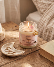 A lit scented candle in a clear glass jar sits on a wooden table beside a small cream dish with rings and an old book. The candle label reads 'Cancer The Heart of the Home' and shows the brand 'Nightgown Scents' and 'BCG Essentials'.
