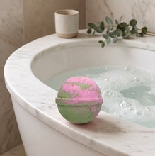 A pastel pink and mint green spherical bath bomb sits on the edge of a white marble bathtub filled with bubbly water. A small ceramic cup and eucalyptus sprig rest on the tub's rim, with a beige tiled bathroom background.