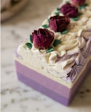 A rectangular layered soap bar with purple and cream sections, decorated with piped cream frosting, dried dark purple roses, and small green leaves, photographed on a light marble-like surface.