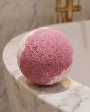 A spherical bath bomb with a marbled pink and white texture, resting on the edge of a white marble bathtub.