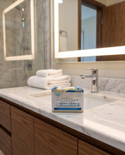 A boxed bar of soap labeled 'Ocean Anchor & Anchor's Anchor' sits on a white marble bathroom counter with a chrome faucet, folded white towels, and a large mirror in the background.