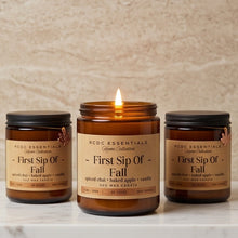 Three amber glass soy wax candles with black lids are arranged on a white marble surface against a beige textured wall. The center candle is lit, and black text at the bottom reads, 'Bring the warmth of autumn to your sanctuary.