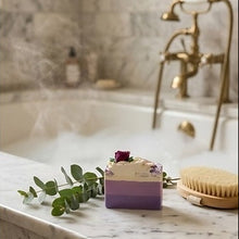 A purple and cream layered bath bar soap with a small rose on top, styled on a marble bathroom counter next to a wooden brush and eucalyptus sprig, with a steamy bathtub and brass faucet in the background.