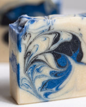 A close-up of a handmade rectangular bar of soap with a marbled swirl pattern of cream, royal blue, and black, resting on a white surface.