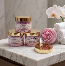 Three jars of Woodstock Handcrafted Soap Scrub arranged on a marble countertop with a pink rose bath bomb, rolled white towels, and white orchids in the background.