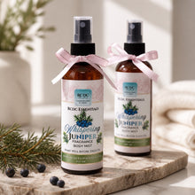 Two bottles of RCDC Essentials Whispering Juniper fragrance body mist on a marble surface with decorative elements.