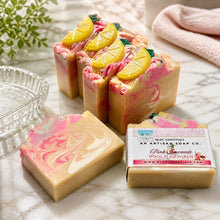 A image of handmade Pink Lemonade soap with pink and yellow colors, decorated with lemon slice designs, displayed with white towel and other decorative items.