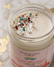 A close-up of a candle in a clear glass jar with a pink label reading 'Signature Zodiac Collection The Grounding Virgo.' The candle wax is creamy off-white, topped with multicolored glitter and confetti, and photographed on a light marble-like surface with gold leaf accents.