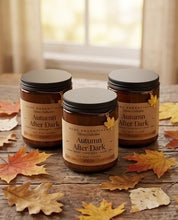 Three amber glass candles with black lids and tan labels reading 'Autumn After Dark' are arranged on a rustic wooden surface with scattered fall leaves. A softly blurred window and curtains appear in the background.