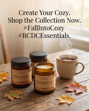 Three amber glass candles with black lids and tan labels are arranged on a rustic wooden table beside a mug of hot cocoa, with scattered autumn leaves. Large text at the top reads, 'Create Your Cozy.