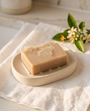 A rectangular bar of pale beige soap with a swirled pattern, resting on a matching ceramic soap dish over a white linen cloth, with small white flowers and green leaves in the background.